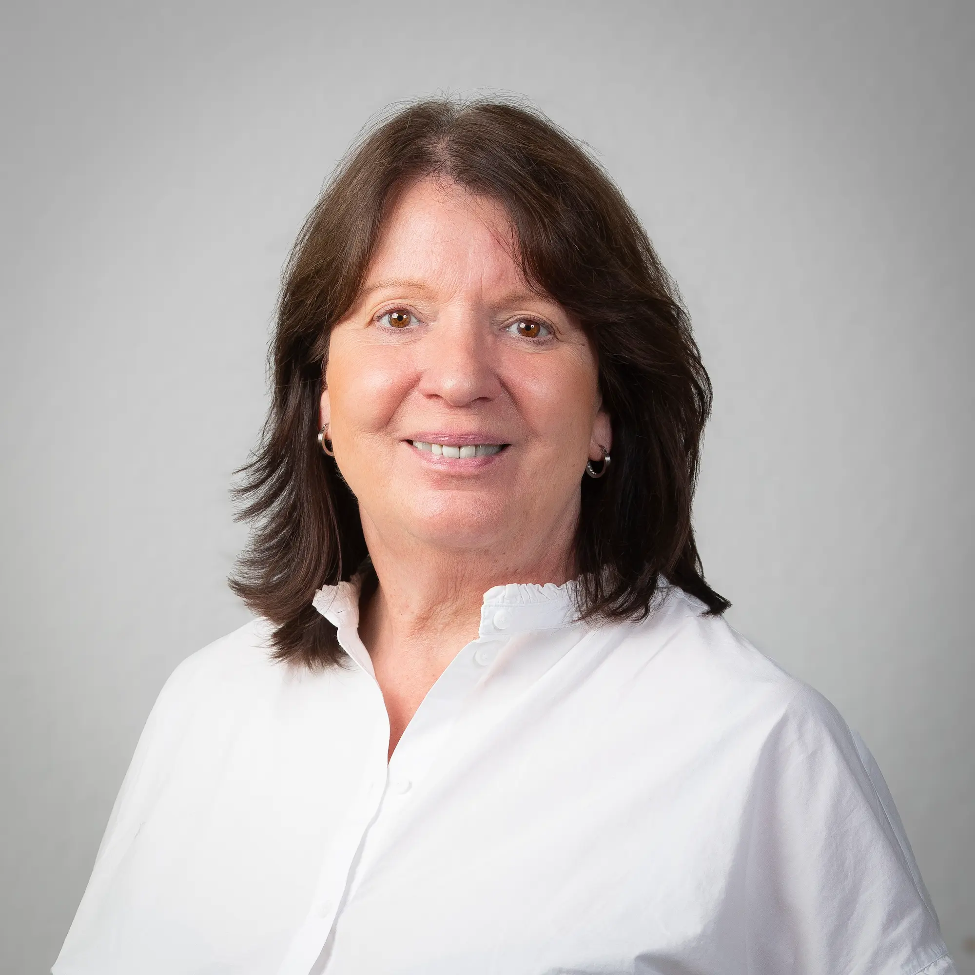 LSB_Mitarbeitende_Annegret_Buchholz Annegret Buchholz mit schulterlangem braunem Haar trägt einen schwarzen Blazer über einem weißen Hemd und lächelt vor einem schlichten hellvioletten Hintergrund.