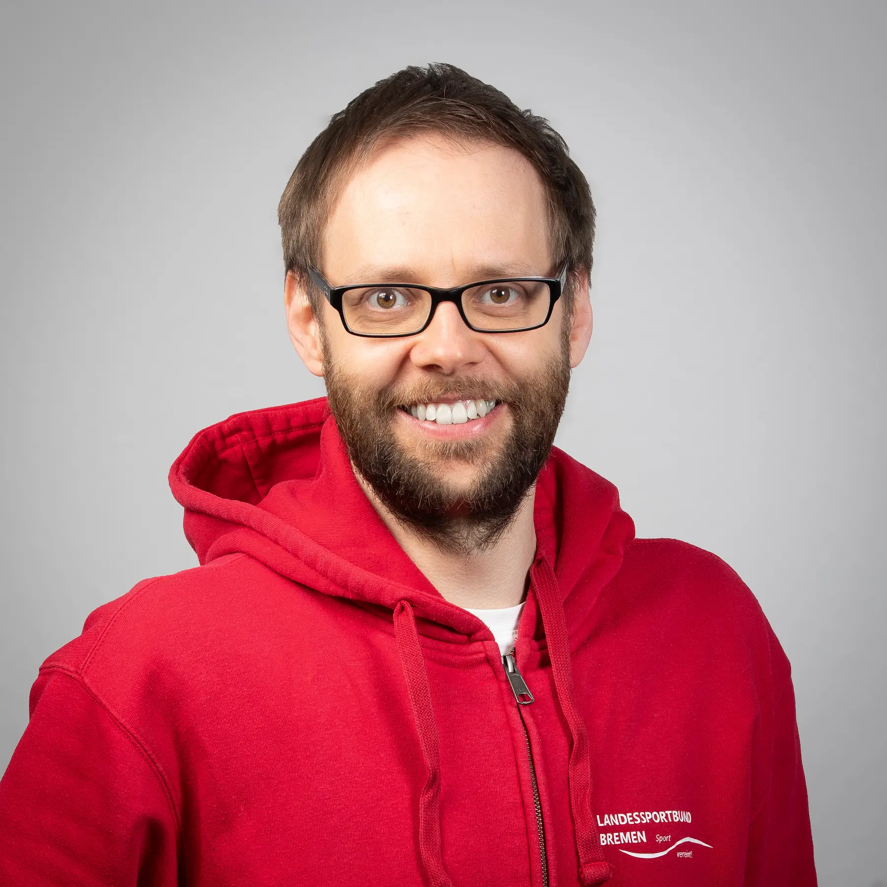 Daniel Fangmann mit kurzen braunen Haaren, Bart und Brille trägt eine schwarze Anzugsjacke über einem weißen Hemd und posiert vor einem schlichten hellblauen Hintergrund.