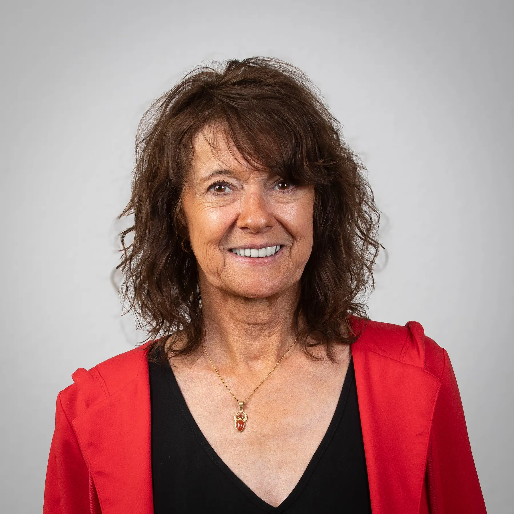 LSB_Mitarbeitende_Gabriele_Vogel Gabriele Vogel mit gewelltem braunem Haar trägt einen schwarzen Blazer, ein weißes Oberteil und mehrere Halsketten und lächelt in die Kamera vor einem schlichten Hintergrund.