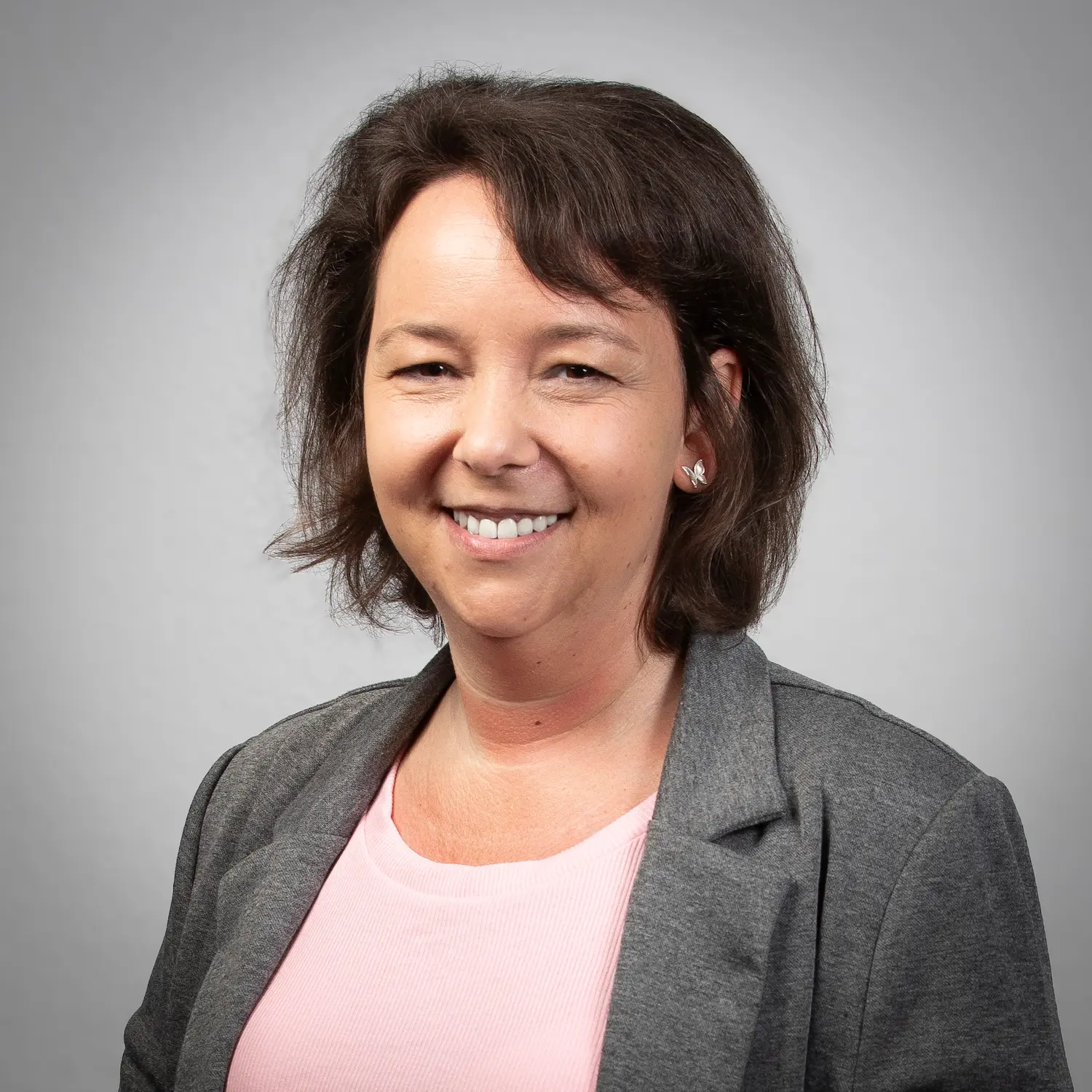 LSB_Mitarbeitende_Jennifer_Nessler Jennifer Neler mit schulterlangem kastanienbraunem Haar, die einen schwarzen Blazer und eine weiße Bluse trägt, lächelt in die Kamera vor einem schlichten hellen Hintergrund.