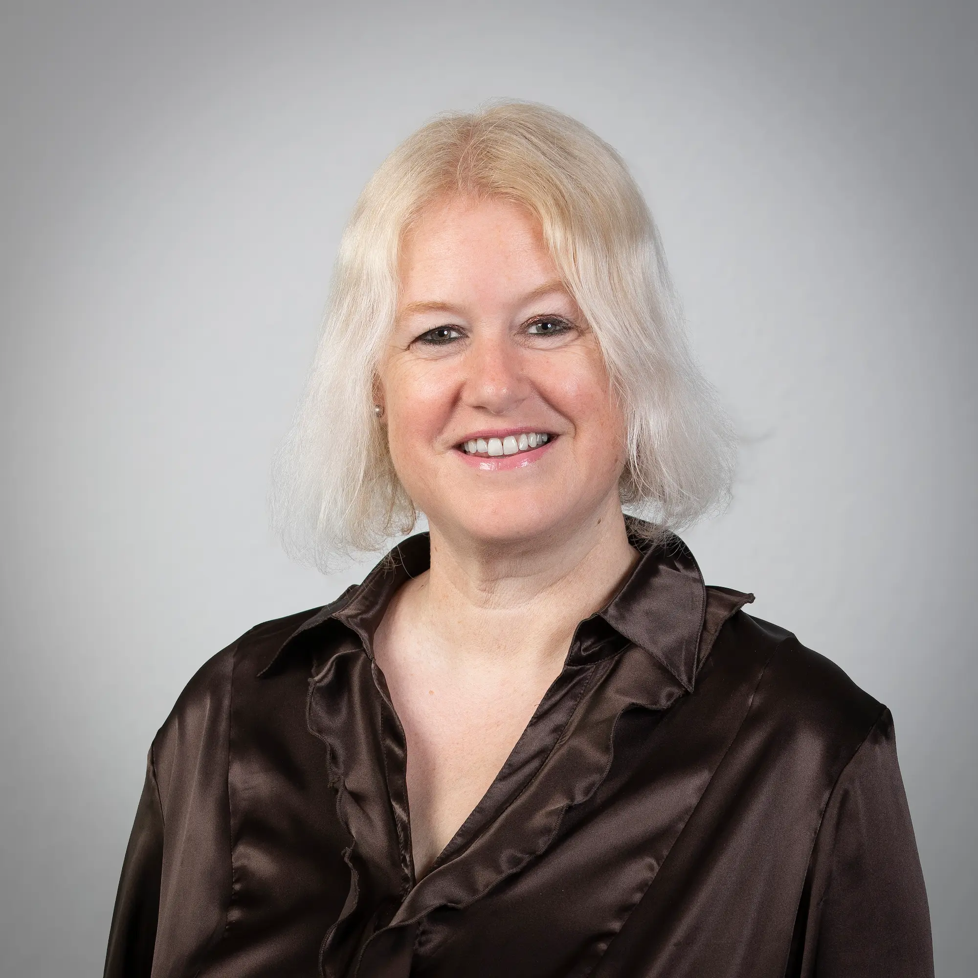 LSB_Mitarbeitende_Tina_Brinkmann Tina Brinkmann-Lange mit kurzen blonden Haaren trägt einen schwarzen Blazer, ein weißes Hemd mit Kragen und einen blaugrünen gemusterten Schal und lächelt vor einem schlichten Hintergrund.