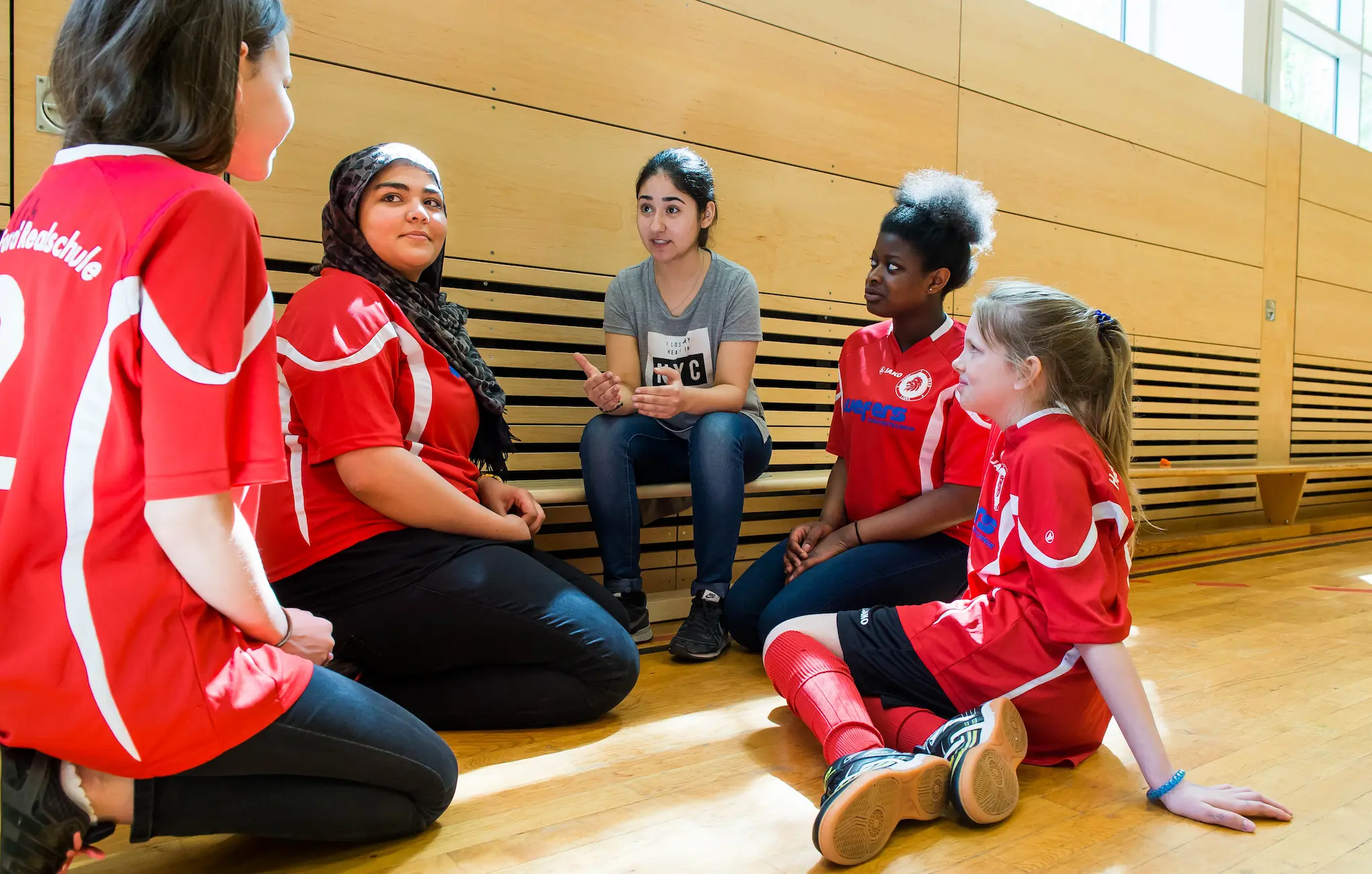 Gruppe mit jungen Mädchen sitzt in der Sporthalle und unterhält sich, alle tragen sportliche Kleidung, ein Mädchen trägt Kopftuch